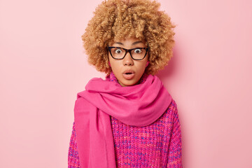 Portrait of surprised shocked woman with blonde curly hair stares through transparent eyeglasses wears knitted jumper and scarf around neck isolated on pink background. Omg and human reactions concept