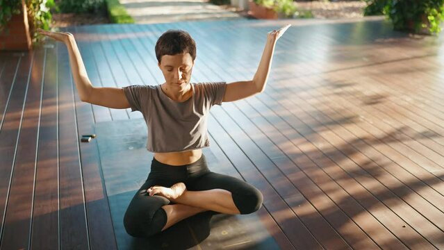 Pleased short-haired woman meditating with closed eyes in the lotus position outdoors