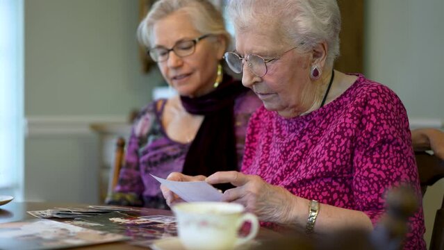 Senior elderly smiling woman looking at old photos and remembering memories with daughter at the dining room table.
