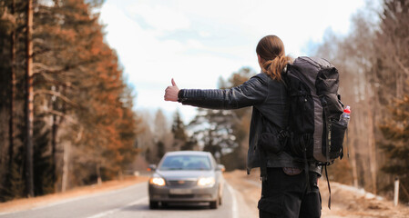 A young man is hitchhiking around the country. The man is trying to catch a passing car for traveling. The man with the backpack went hitchhiking to the south.