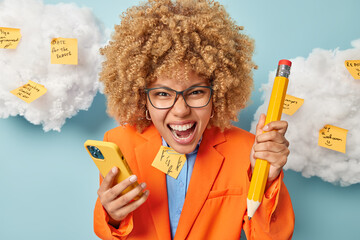 Annoyed curly haired woman shouts angrily dressed in formal clothes holds big pencil and smartphone works in office poses against blue background white coluds with memo stickers written things to do