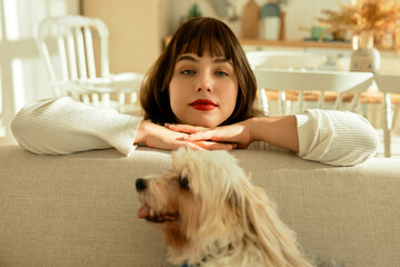 Attractive confident girl of 20s with brown hair, wearing red lipstick resting head on folded palms leaning on back of sofa, looking at camera, while her cute pet dog sitting in front. Selective focus