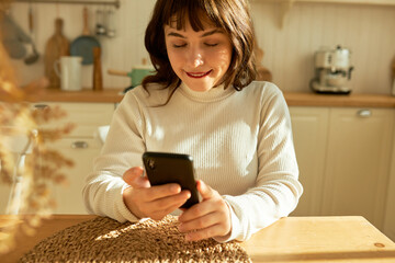 Flirty attractive brunette girl of 20s wearing red lipstick using smartphone sitting at kitchen table, biting her lower lip chatting with guys in dating phone application, searching for real love