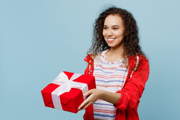 Side view young woman of African American ethnicity wears red jacket hold giving present box with gift ribbon bow isolated on plain pastel light blue cyan background Wet fall weather season concept.