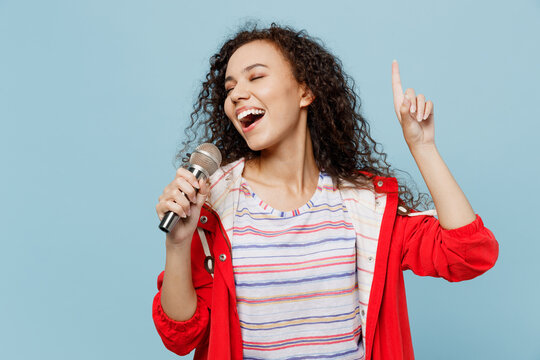 Young Singer Woman Of African American Ethnicity 20s She Wear Red Jacket Sing Song In Microphone Point Finger Up Isolated On Plain Pastel Light Blue Cyan Background. Wet Fall Weather Season Concept.