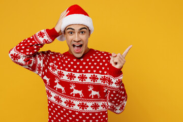 Merry young man wear knitted christmas sweater Santa hat posing point index finger aside indicate...