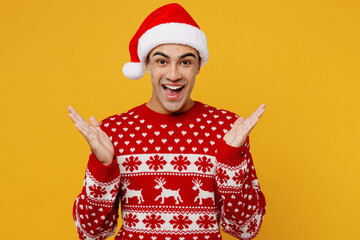 Merry shocked excited young man wear red knitted christmas sweater Santa hat posing spread hand look camera say wow isolated on plain yellow background Happy New Year 2023 celebration holiday concept