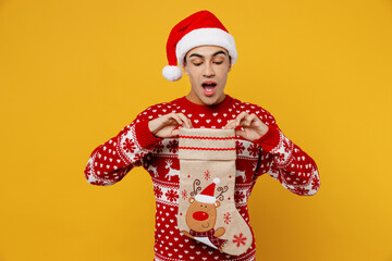 Merry surprised fun young man wear red knitted Christmas sweater Santa hat posing hold stocking sock look camera isolated on plain yellow background. Happy New Year 2023 celebration holiday concept.