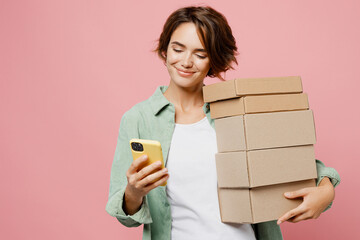 Young happy woman wear green shirt white t-shirt hold hand brown clear blank craft stack cardboard boxes mock up use mobile cell phone isolated on plain pastel light pink background studio portrait