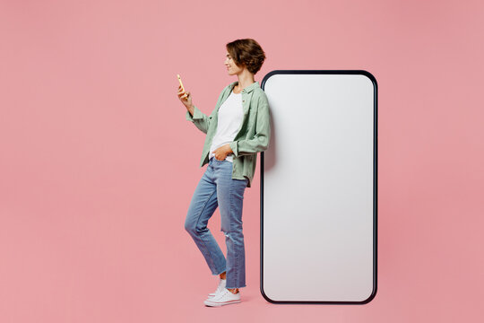 Full body side view young woman wear green shirt white t-shirt near big huge blank screen mobile cell phone smartphone with mockup use mobile cell phone isolated on plain pastel light pink background.