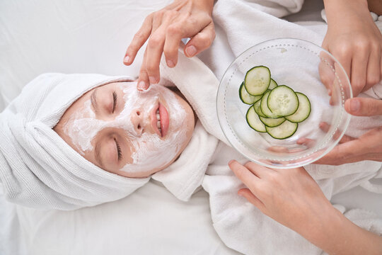 Top View Of Crop Anonymous Mother Applying Facial Mask On Kid In Bathrobe With Towel On Head Relaxing On Soft Bed With Closed Eyes