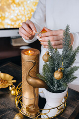 Unrecognizable woman featuring fire at big golden candle.Small minimalistic fir tree, festive glittering balls decorations on wooden tray. Christmas holiday celebration. New Year Xmas home hygge decor
