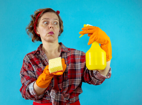 Pin-up woman in a plaid shirt holding a yellow spray bottle and a washcloth for washing on an isolated blue background. portrait woman american pin-up. Woman cleaning service in orange gloves