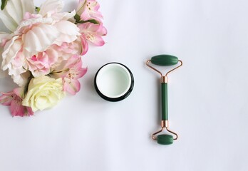Stone facial massager made of green jade, , a delicate bouquet and a jar of cosmetic cream on a white background. View from above. Face care products. View from above. Everything is for beauty.