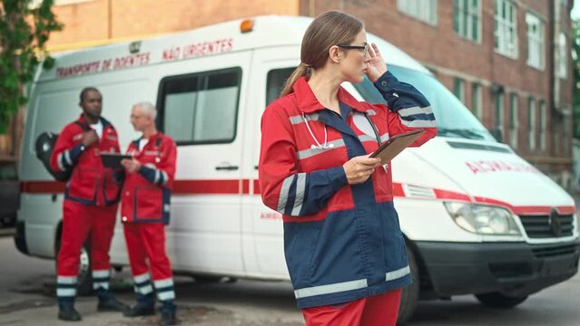Young beautiful Caucasian female doctor in glasses texting on tablet device while standing outdoor near ambulance car. Male paramedics nurses in red uniform on background, first aid, emergency concept