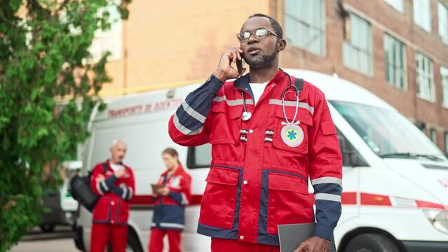 Camera moving back from African American professional male doctor in glasses talking on smartphone standing outdoor. Paramedics nurses in red uniform on background near ambulance car