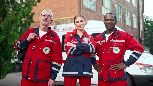 Portrait of team of paramedics in red medical uniform posing outdoors. Male and female doctors standing in street near ambulance and smiling at camera. Medical workers. Professional nurses, first aid