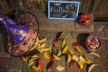 Top view: a blackboard with the inscription Halloween next to a pumpkin in wizard's hat on a wooden threshold with fallen yellow and red leaves in autumn. Porch or backyard decoration for Halloween