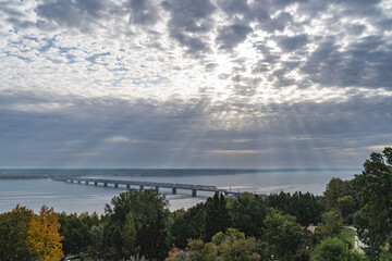 Fototapeta na wymiar Real sun rays through the clouds in the sky the bridge across the Volga river in Ulyanovsk, built under the tsar in Russia. Operating iron bridge. Connects two shores of one city.