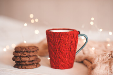 Cup of coffee with red knit cloth textile with chocolate biscuits in stack over Christmas lights close up in bed