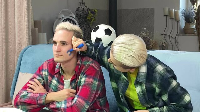Young stylish white couple preparing to cheer Argentina soccer team on championship at home. Happy young woman painting Argentine flag on male face indoors. Support Argentina football at world cup
