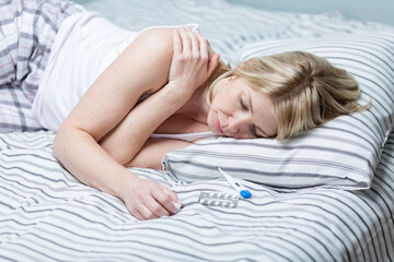 A young woman lies on a bed with a thermometer and pills. Blonde in pajamas feeling unwell....