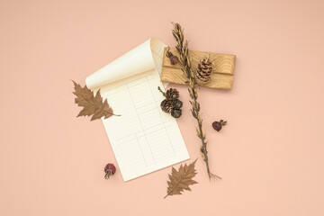 Autumn composition of white notepad with blank sheets among yellowed leaves and pinecones. Decorative arrangement on light pink background