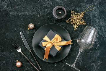 Stylish table setting, gift box, star and balls with black plate, cutlery and wineglass on black background. Christmas concept. Top view, flat lay