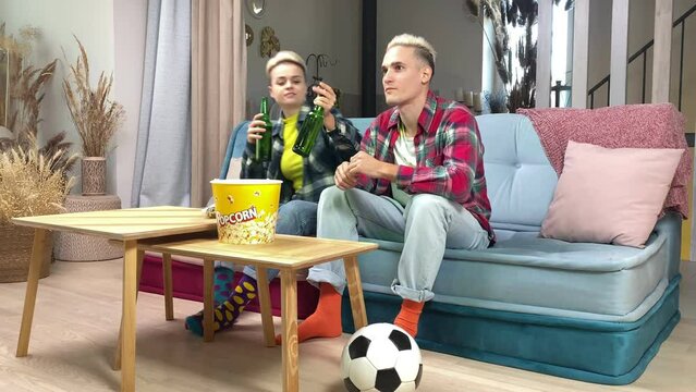 Cheerful young spouses watching live soccer match on TV at home. Enthusiastic husband cheering sport team while wife bringing beer indoors. Football fan couple having pastime together, watching game