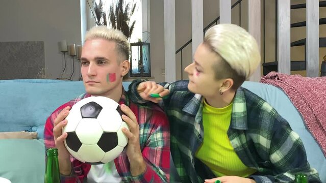 Young stylish white couple preparing to cheer Portugal soccer team on championship at home. Happy young woman painting Portuguese flag on male face indoors. Support Portugal football at world cup