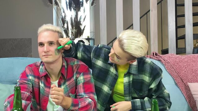 Young stylish white couple preparing to cheer Brazil soccer team on championship at home. Happy young woman painting Brazilian flag on male face indoors. Support Brazil football at world cup