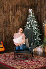 Obraz premium Beautiful stylish woman in a pink sweater and blue jeans sits near on a chair and a Christmas tree. Christmas background. Concept: Merry Christmas, noel, santa claus, family holidays