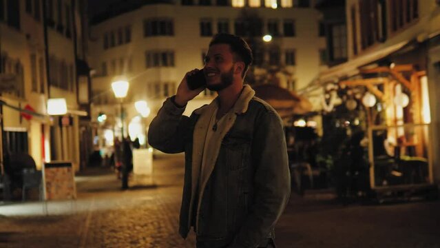 Funny positive cheerful man standing in the evening or night and chatting on phone with girlfriend, laughing, having a good mood. Male talking on phone with friend, cell phone outdoors lights