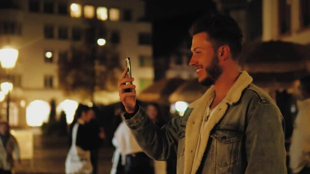 Cheerful man having video conference call online, using mobile phone smartphone. Young male congratulate family or friends with Christmas or New Year on the street with lights, illuminated old town 