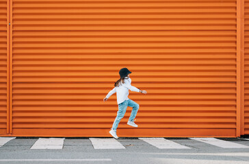 Little fashion-dressed girl funny jumping crossing a pedestrian zebra crosswalk on the orange wall background. Urban people living and a street everyday life concept image.