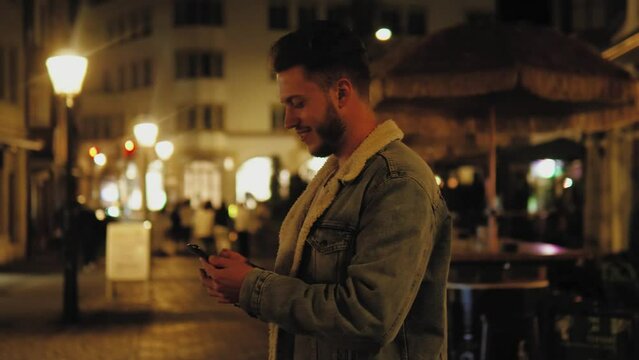 Cheerful caucasian man looking at phone screen, browsing social media, scrolling internet at night at cozy illuminated street. Christmas atmosphere. Old town in lights