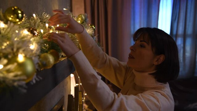Brunette caucasian woman in white sweater dress adjusting Christmas lights on garland with golden Christmas baubles. Real time video. Selective focus. Winter holidays mood theme.