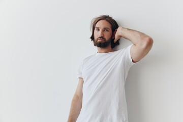 A man in a white T-shirt and blue jeans stands against a white wall and poses