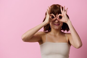 a funny, playful woman with short hair poses standing on a pink background showing the OK sign with her fingers near her eyes, covering them with her hands. Horizontal photo with empty space 