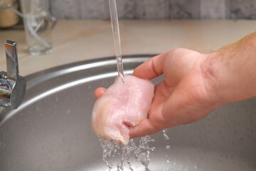Fototapeta premium A man washes defrosted raw chicken fillet. Fresh raw chicken breast for cooking chopped chicken cutlets. A man washes a chicken fillet under a stream of water for home cooking.