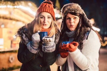 Two women with hot mulled wine in cups in front of Christmas market stand