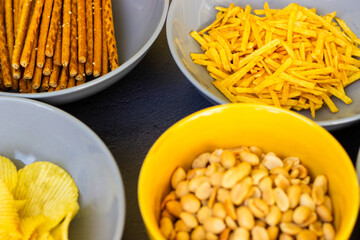Salty snacks assortment shot on dark slate table. The picture includes potato chips, popcorn, peanut, cheese sticks, ketchup sticks, tzatziki chips, smoky flips, and others