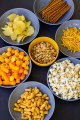 Salty snacks assortment shot on dark slate table. The picture includes potato chips, popcorn, peanut, cheese sticks, ketchup sticks, tzatziki chips, smoky flips, and others
