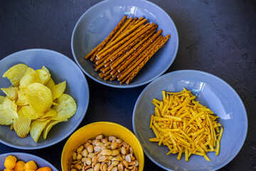 Salty snacks assortment shot on dark slate table. The picture includes potato chips, popcorn, peanut, cheese sticks, ketchup sticks, tzatziki chips, smoky flips, and others