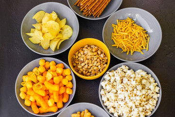 Salty snacks assortment shot on dark slate table. The picture includes potato chips, popcorn, peanut, cheese sticks, ketchup sticks, tzatziki chips, smoky flips, and others
