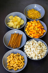 Salty snacks assortment shot on dark slate table. The picture includes potato chips, popcorn, peanut, cheese sticks, ketchup sticks, tzatziki chips, smoky flips, and others