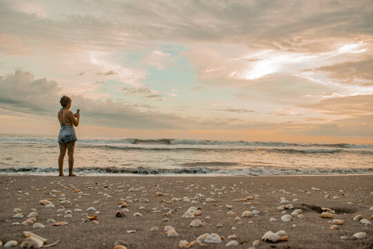  Mujer a la orilla del mar observando el atardecer en El Salvador  mientras toma fotos con su celular 