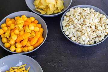 Salty snacks assortment shot on dark slate table. The picture includes potato chips, popcorn, peanut, cheese sticks, ketchup sticks, tzatziki chips, smoky flips, and others