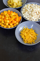 Salty snacks assortment shot on dark slate table. The picture includes potato chips, popcorn, peanut, cheese sticks, ketchup sticks, tzatziki chips, smoky flips, and others