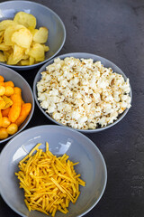 Salty snacks assortment shot on dark slate table. The picture includes potato chips, popcorn, peanut, cheese sticks, ketchup sticks, tzatziki chips, smoky flips, and others